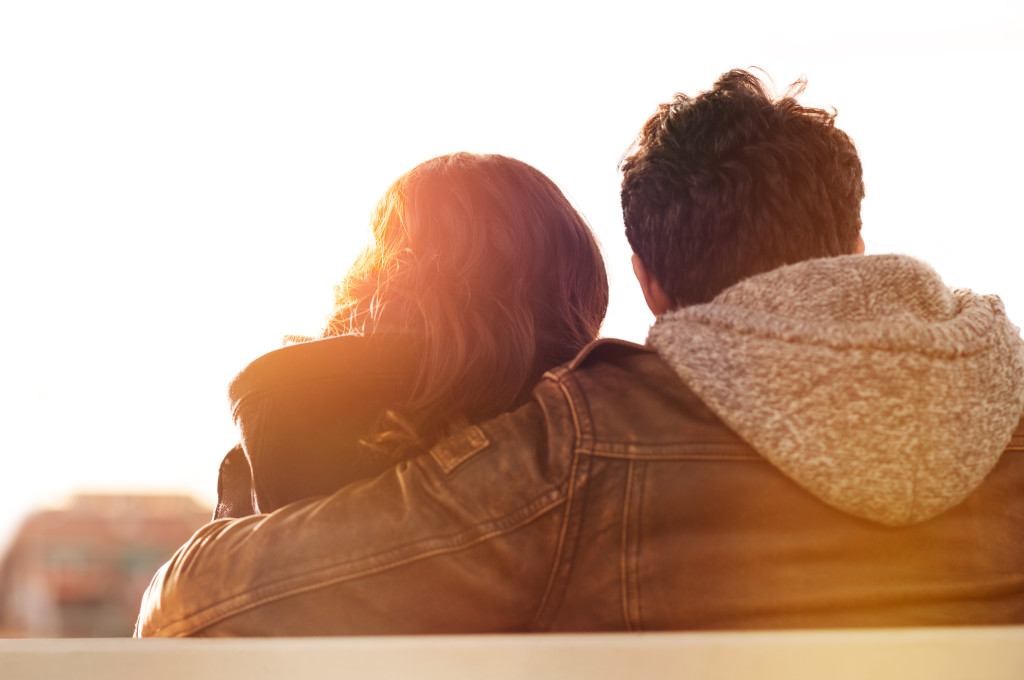 Closeup rearview of man embracing his woman while sitting on bench. They are sitting outdoor and looking away in a beautiful sunset light. Contemplative and aspirational mood.