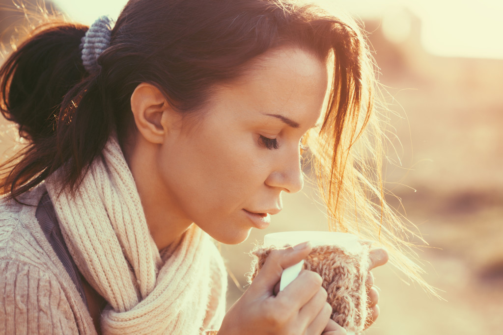 Woman wearing warm knit clothes drinking cup of hot tea or coffee outdoors in sunlight